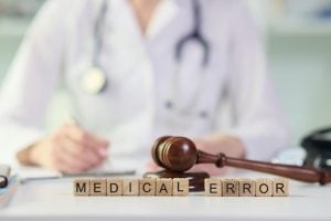 A doctor sits at a desk with a stethoscope, gavel, and wooden blocks spelling "MEDICAL ERROR" in front of them.