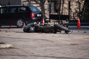 A damaged motorcycle lies on its side in the road near a black car and orange traffic cones, indicating a recent accident.