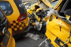 Several yellow taxis appear severely damaged in a collision, with crumpled and broken car parts visible in close proximity on a gravel surface.
