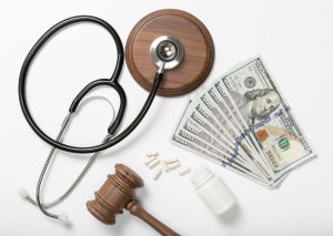 A stethoscope, a wooden gavel, a bottle of pills, and several hundred-dollar bills are arranged on a white surface.