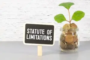 A small sign reading "Statute of Limitations" stands next to a glass jar filled with coins and a plant inside, against a white brick wall background.