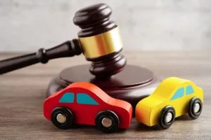 A judge’s gavel is shown behind two toy cars, one red and one yellow, on a wooden surface, representing legal issues related to automobiles.