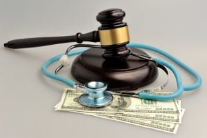 A gavel and stethoscope rest on top of several one-hundred-dollar bills, symbolizing the intersection of healthcare, law, and financial matters.