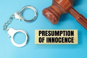 A wooden gavel, metal handcuffs, and a block labeled "PRESUMPTION OF INNOCENCE" are displayed on a blue background.