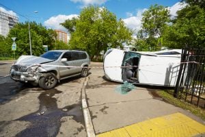Two cars involved in an accident at an intersection; one vehicle is overturned on its side, and the other has a damaged front end. Broken glass and police tape are visible.