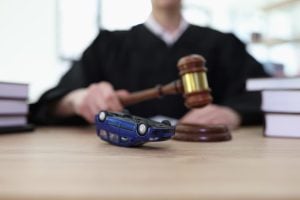 A judge in black robes holds a gavel near a model car overturned on a desk, with books stacked on both sides, suggesting a legal case involving a car accident.