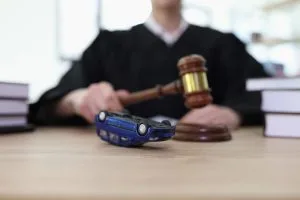 A judge in black robes holds a gavel near a model car overturned on a desk, with books stacked on both sides, suggesting a legal case involving a car accident.