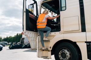 A person wearing a reflective orange vest climbs into the cab of a white truck parked in a lot with several tires and cars visible in the background.