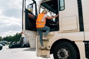 A person wearing a reflective orange vest climbs into the cab of a white truck parked in a lot with several tires and cars visible in the background.