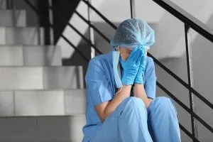 A healthcare worker in scrubs, gloves, and a hair cover sits on stairs with face in hands, appearing distressed or exhausted.