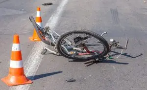 A damaged bicycle lies on its side on an asphalt road, surrounded by three orange and white traffic cones.