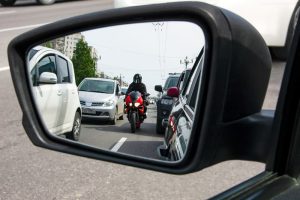 A motorcycle and several cars are visible in traffic through a car’s side mirror.