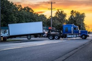 A tow truck is pulling a white box truck on a road at sunset, with power lines and trees in the background.