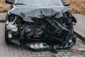 A black car with severe front-end damage, including a crumpled hood, exposed engine parts, and detached headlights, parked on a paved surface.