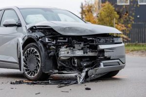 A silver car with significant front-end damage and deployed airbags is parked on a road after a collision.