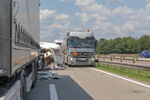 Two large trucks and a white camper involved in a collision on a highway, with debris scattered across the road and traffic stopped.