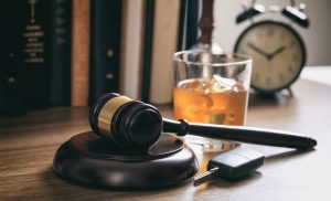 A judge’s gavel, a glass of whiskey, and car keys on a desk, with books and an alarm clock in the background.