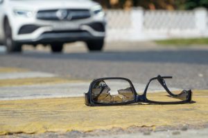 A pair of broken eyeglasses lies on a crosswalk, with a white car approaching in the blurred background.