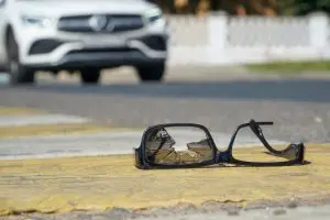 A pair of broken eyeglasses lies on a crosswalk, with a white car approaching in the blurred background.