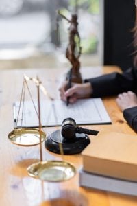 A person signs a document at a desk with a gavel, scales of justice, books, and a Lady Justice statue.