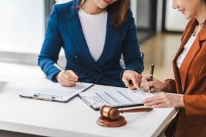 Two women sitting at a desk, reviewing and signing documents with a gavel placed on the table beside them.