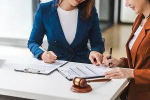 Two women sitting at a desk, reviewing and signing documents with a gavel placed on the table beside them.