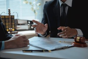Two people in business suits sit at a desk, discussing a contract document. A gavel, pen, and legal scales are visible in the background.