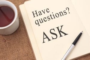 Open notebook with the words "Have questions? ASK" written on a page, next to a pen and a cup of tea on a burlap surface.