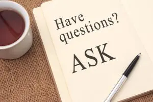 Open notebook with the words "Have questions? ASK" written on a page, next to a pen and a cup of tea on a burlap surface.
