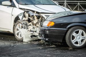 Two cars, one white and one black, have collided head-on, causing significant damage to the front ends of both vehicles on a wet road.