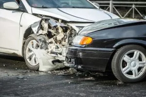 Two cars, one white and one black, have collided head-on, causing significant damage to the front ends of both vehicles on a wet road.