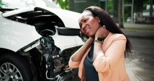 A woman stands in front of a damaged car, holding her neck and appearing to be in pain, possibly from a recent accident.