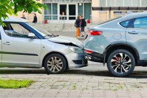 A silver car has rear-ended a gray SUV on a city street, with visible damage to the front of the car and the rear of the SUV. People walk in the background.