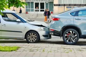A silver car has rear-ended a gray SUV on a city street, with visible damage to the front of the car and the rear of the SUV. People walk in the background.