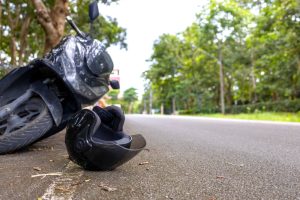 A motorcycle is lying on its side near the edge of a road, with a black helmet resting on the pavement in the foreground. Trees line both sides of the road.