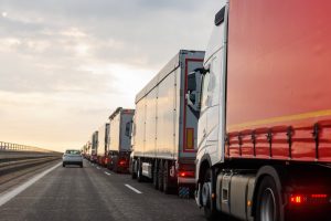 A row of large trucks and a car drive along a multi-lane highway under a cloudy sky.