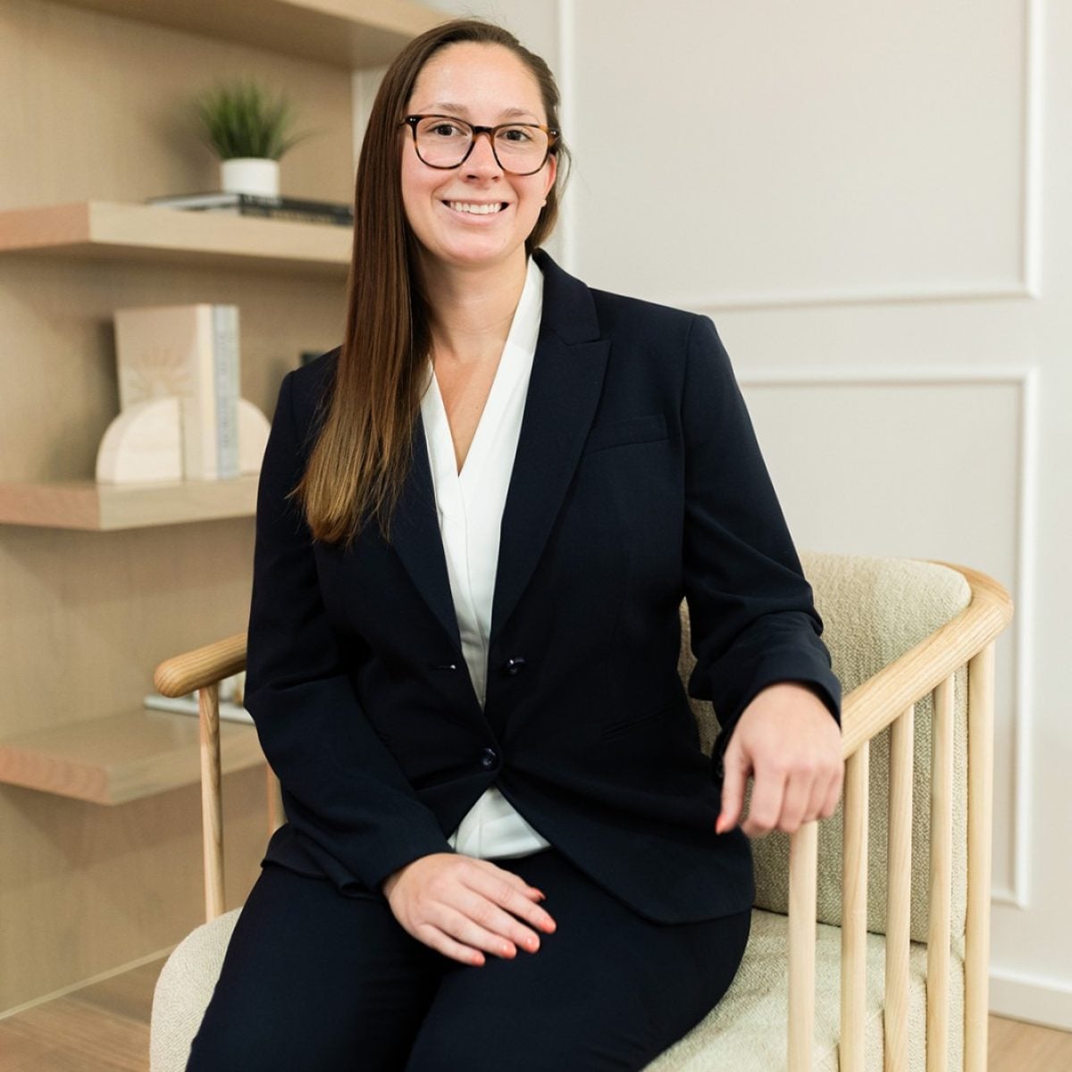 A woman in a dark suit and glasses sits on a chair in a modern office space with bookshelves in the background.