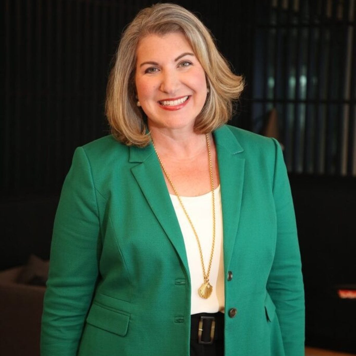 Woman with shoulder-length blonde hair smiles and stands indoors, wearing a green blazer, white top, black pants, and a long gold necklace.