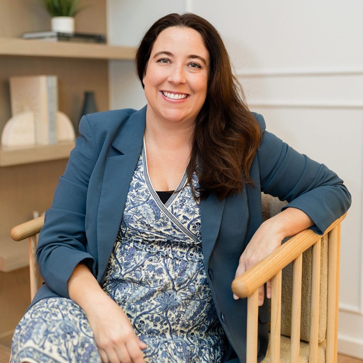 A woman with long brown hair, wearing a blue patterned dress and blue blazer, sits and smiles in a wooden chair in a modern, well-lit room with shelves behind her.