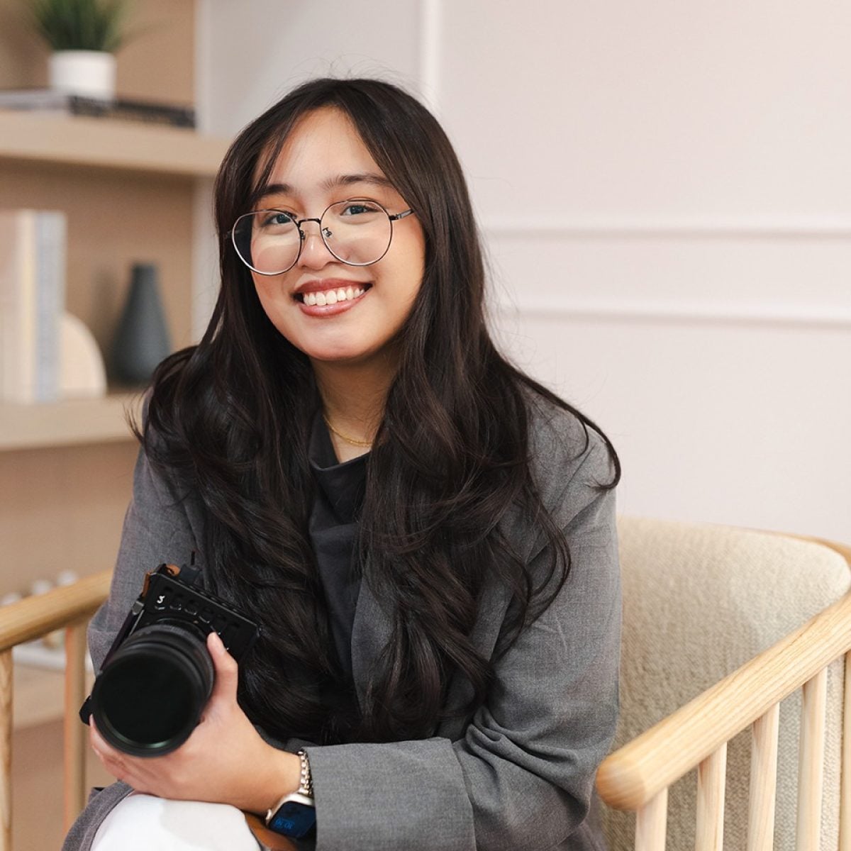 A woman with long dark hair and glasses sits on a chair, smiling, and holding a camera. She is wearing a gray blazer and white pants. Shelves with books and a plant are in the background.