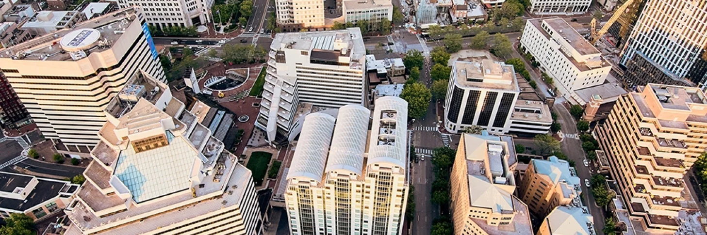 Aerial view of a downtown area with multiple modern office buildings, tree-lined streets, and a sports field in the background.